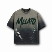 A short-sleeve crewneck t-shirt with a dark gray to black gradient wash, featuring a large green spray-paint style 'MELATO' graphic on the chest, smaller 'MELATO STUDIOS' text, and white paint splatters near the lower front.